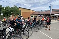 Gruppe von Radfahrern mit Fahrrädern steht auf einem Parkplatz vor einem große gelb-orangem Gebäude unter bewölktem Himmel.