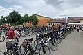 Viele geparkte Fahrräder auf einem großen Parkplatz vor einem großen gelb-orangem Gebäude mit Menschen im Hintergrund bei bewölktem Himmel.