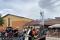Stadtpfarrer segnet Fahrräder mit Weihwasser, neben ihm eine weitere Person mit einem Behälter mit Weihwasser, umgeben von Menschen und Fahrrädern vor einem großen gelb-orangen Gebäude im Hintergrund.