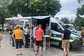 Menschen, darunter ein Polizeimitarbeiter stehen an einem Polizeifahrzeug mit ausgefahrener Markise auf einem Parkplatz mit Bäumen und weiteren Autos im Hintergrund.
