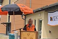 Mann in orange-schwarzem Shirt spricht am Rednerpult aus Holz mit 9 unterschiedlichen Wappen an der Front auf einem gepflastertem Platz, unter orange-schwarzem Kolping-Sonnenschirm. Im Hintergrund großes hohes Gebäude in orange/gelb und ein Werbebanner mit der Aufschrift Kolping.