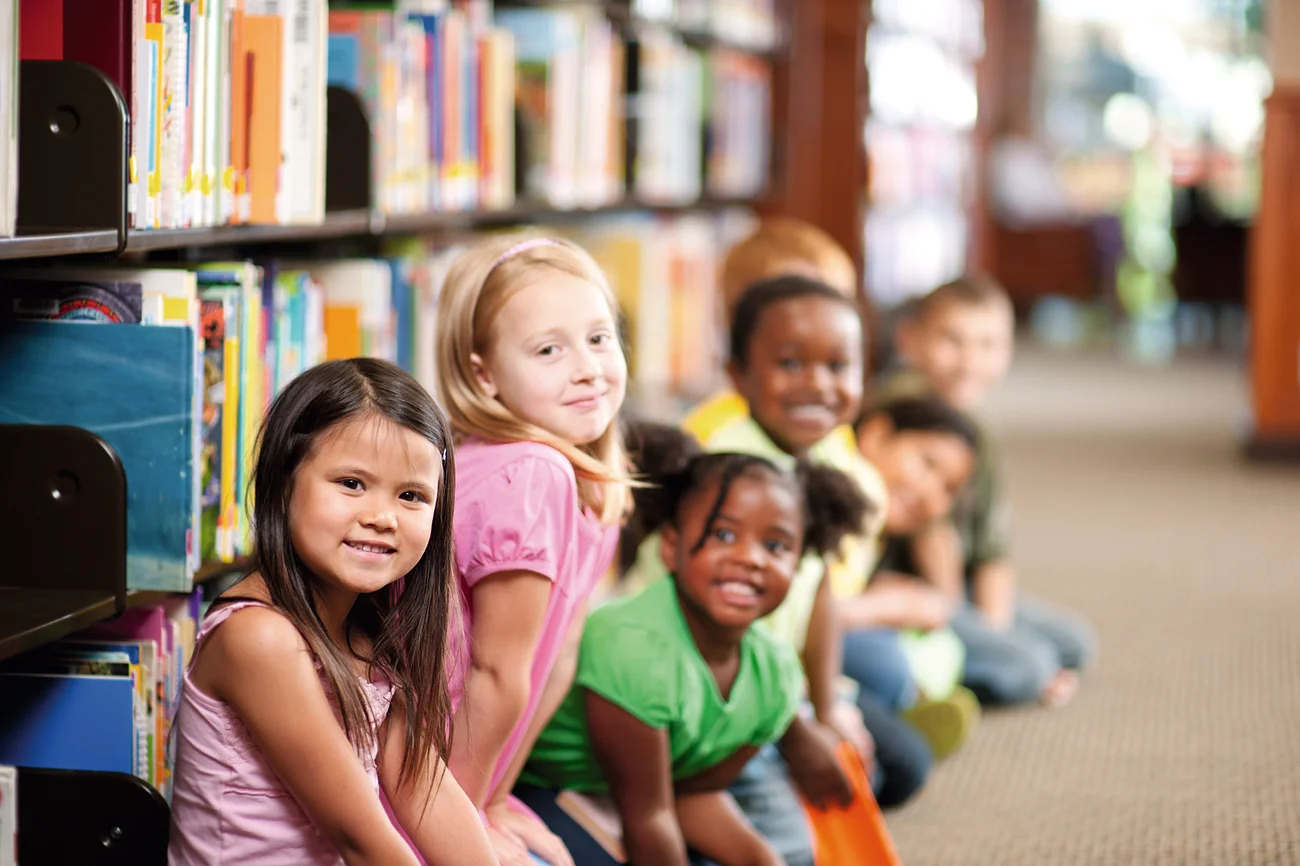 Kinder sitzen in einer Reihe auf dem Boden einer Bibliothek, Bücherregale im Hintergrund.