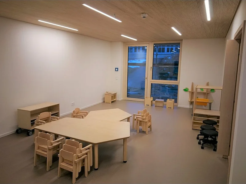 Kita-Gruppenraum in Neubau Kindergarten Monheim Gruppenraum im städtischen Kindergarten Monheim mit Kindertischen und Stühlen aus Holz, einem Fenster und verschiedenen Regalen.
