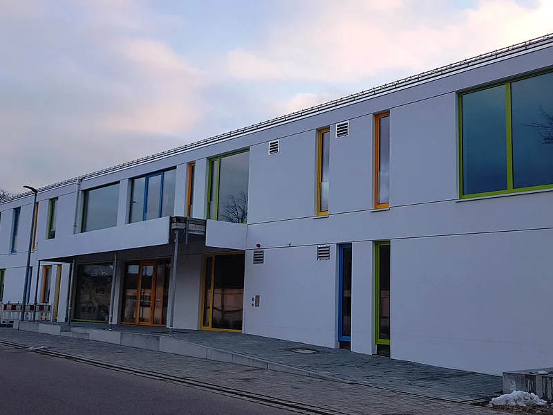 Neuer Kindergarten Monheim Außenansicht Modernes zweistöckiges Gebäude, der städtische Kindergarten Monheim in weiß, mit vielen bunten Fensterrahmen an einer Straße bei bewölktem Himmel.