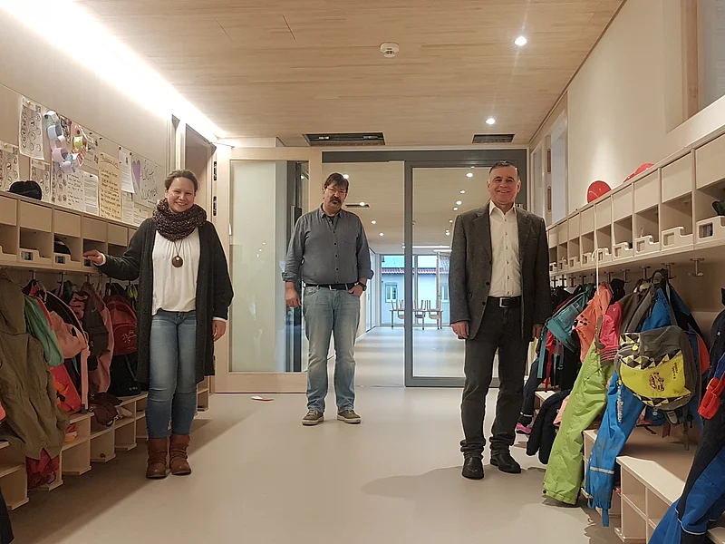 Über den neuen Kindergarten freuen sich die Leiterin Susanne Utjesinovic, Stadtbaumeister Richard Meyer und Bürgermeister Günther Pfefferer (v.l.n.r) Drei Personen stehen in einem Flur mit Kindergarderoben und bunten Jacken. Im Hintergrund ist ein Raum mit Tischen und kleinen Stühlen sichtbar.