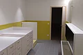 Sanitärbereich im Neubau Kindergarten Monheim Sanitärbereich im Kindergarten Monheim: ein Raum mit weißen breiten Schränken mit Ablagefläche, gelb-grünen Wandfliesen bis zur Hälfte der Wände, grauem Boden und eine offener Tür zum nächsten Raum.
