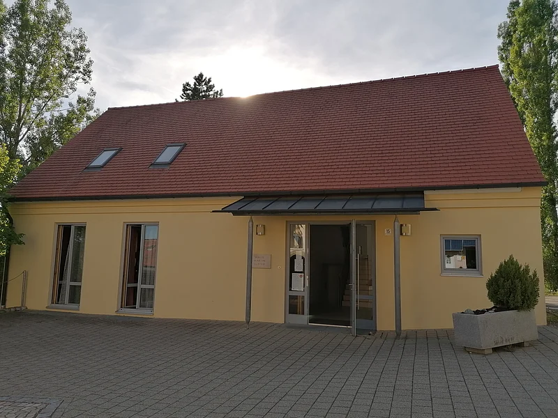 Haus Martin Luther in Monheim - Evangelisches Gemeindehaus Das Haus Martin Luther in Monheim, ein evangelisches Gemeindehaus: Ein gelbes Gebäude mit rotem Satteldach, zwei Fenstern und einer offenen Eingangstür, umgeben von Bäumen. Vor dem Haus steht ein bepflanztes Beet.