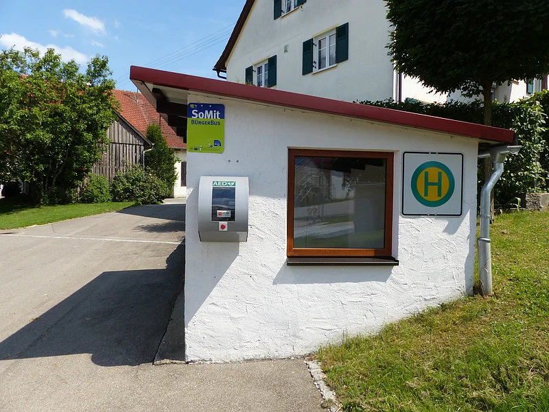 Kleine weiße Bushaltestelle mit rotem Dach, einem Fenster und einem Defibrillator mit der Aufschrift "AED" an der Wand. Auf dem Bushäuschen ein Haltestellenschild und ein grün-blaues Schild mit der Aufschrift "SoMit Bürgerbus" angebracht. Ländlichen Umfeld.