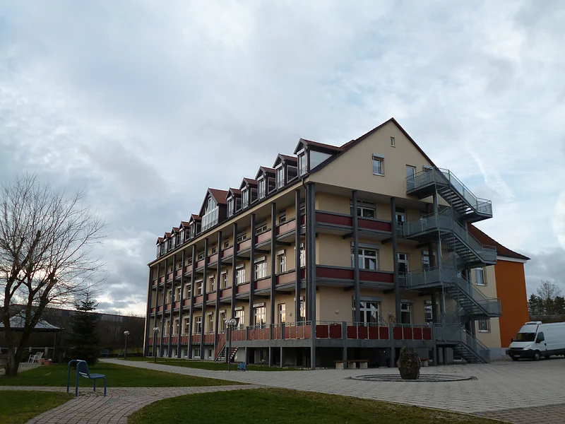 Donau-Ries Seniorenheim Monheim Mehrstöckiges orange gestrichenes Gebäude mit vielen Fenstern, Balkonen und einer großen Außentreppe, umgeben von Gehwegen und Rasenfläche. Vorderhalb ein gepflasterter Platz mit einem großen Stein drauf. Links im Bild eine Sitzbank. Im Hintergrund bewölkter Himmel.