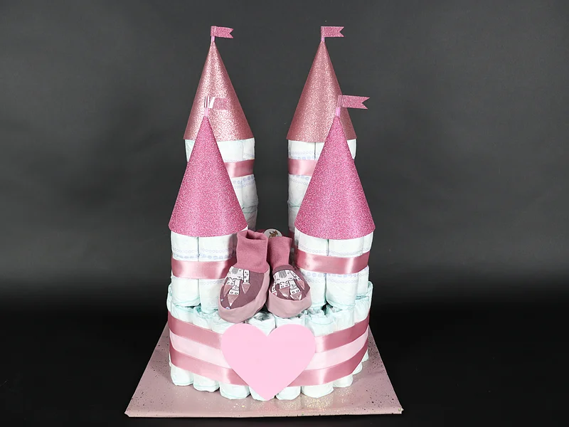 Windelrella Windeltorten - Windeltorte Prinzessinnenschloss Windeltorte in Form eines Schlosses mit rosa Türmen und Babyschuhen auf schwarzem Hintergrund.