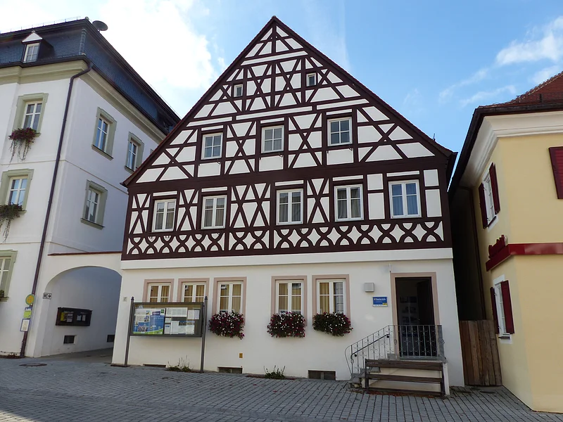 Tourist-Information der Stadt Monheim/Monheimer Alb: ein Fachwerkhaus mit drei Stockwerken, weißen Wänden und braunen Balken. Davor steht eine Sitzbank und ein Schaukasten auf einem gepflasterten Platz. Links davon ein weißes dreistöckiges Gebäude. Rechts davon ein gelbes zweistöckiges Gebäude mit roten Fensterläden.