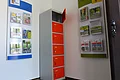 Ein orange-grauer Indoor-Ladeschrank für eBike-Akkus mit insgesamt sechs Fächern und einem offenen Fach steht zwischen zwei Wanddisplays mit Broschüren in dem Vorraum der Touristinformation Monheim.