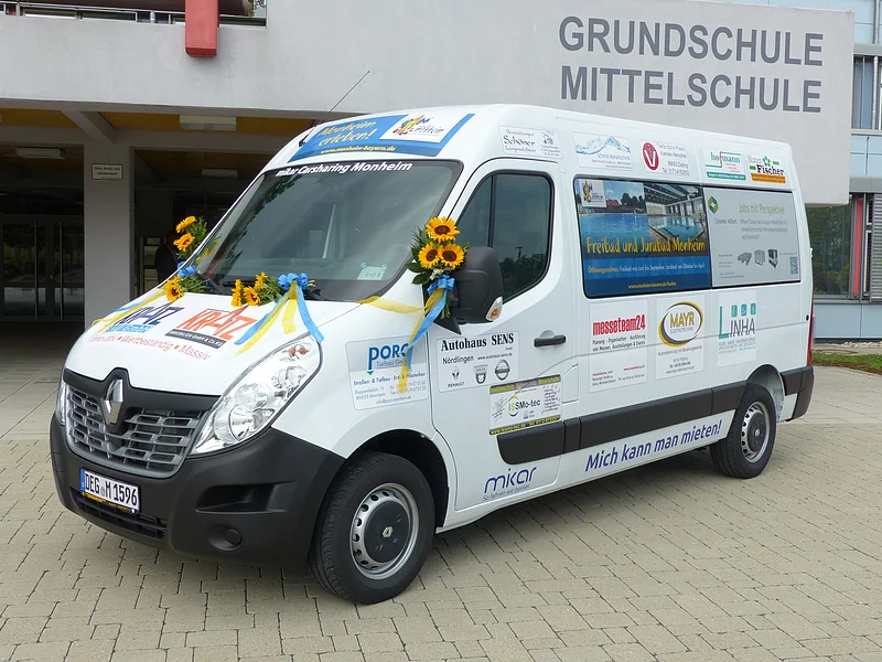 Ein weißer Transporter vom Car-Sharing-Anbieter Mikar mit Werbeaufdrucken und Sonnenblumen steht vor einem Gebäude mit der Aufschrift "Grundschule Mittelschule" in Monheim.