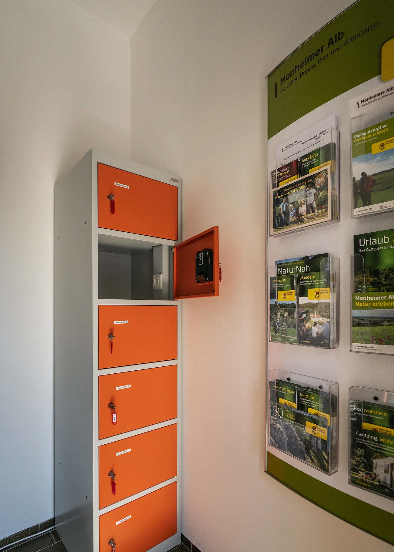 Ein orange-grauer Indoor-Ladeschrank für eBike-Akkus mit insgesamt sechs Fächern und einem offenen Fach steht neben einem Wanddisplay mit Broschüren in dem Vorraum der Touristinformation Monheim.