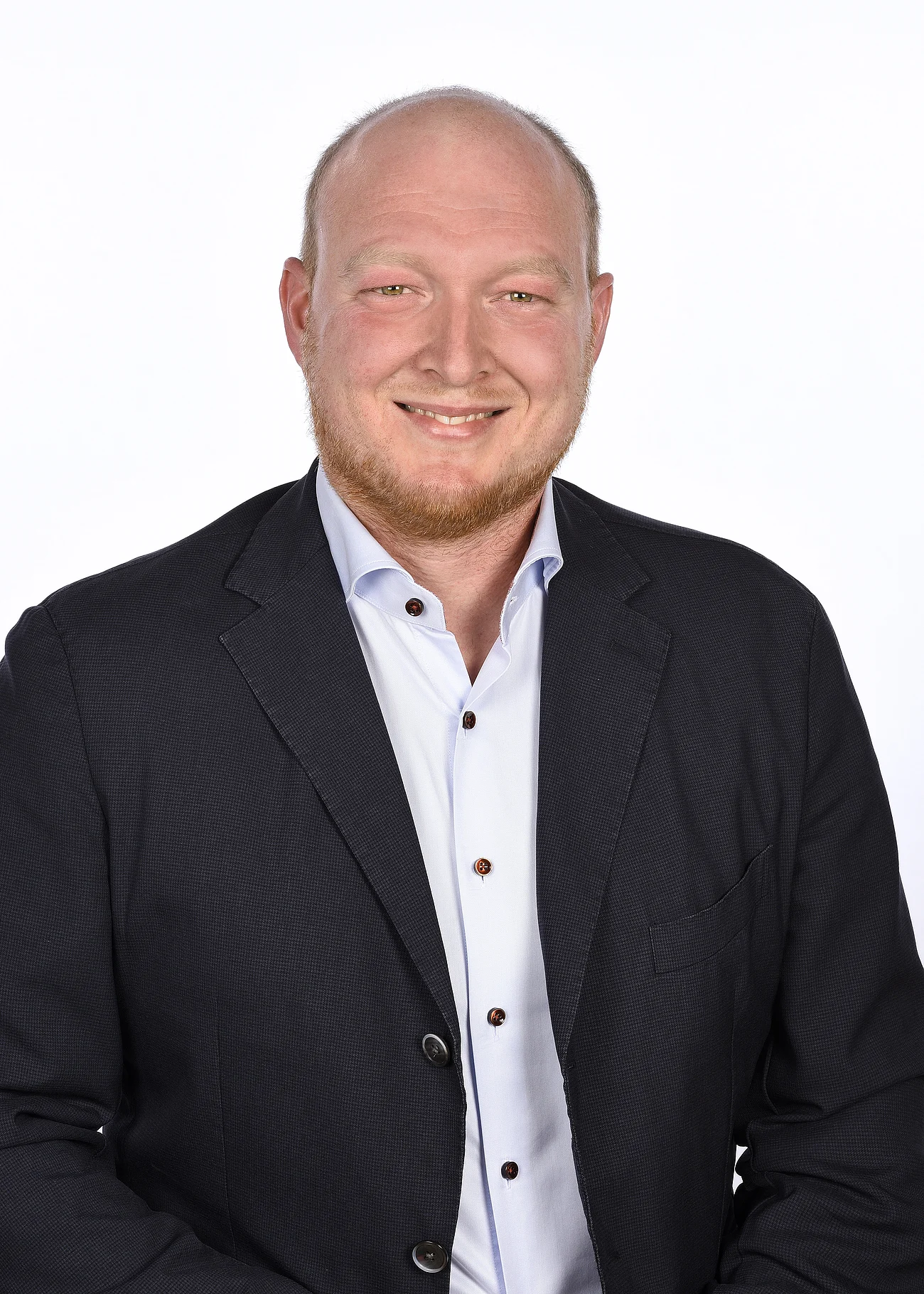 Ein Mann in einem schwarzen Anzug und Hemd lächelnd vor einem weißen Hintergrund.