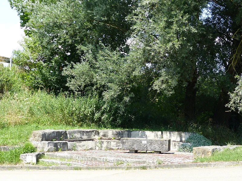 Rastplatz für Radler in Itzing Rastplatz für Radler in Form von einer steinernen Sitzbank aus Stufen vor dichten grünen Bäumen und Sträuchern im Freien bei Tageslicht. Im Vordergrund ein Stück Straße zu sehen.