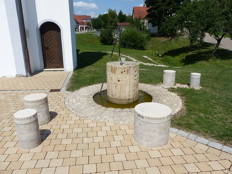 Brunnen mit zylindrischem Steinbecken mit laufendem Wasser und einem gepflastertem Rundplatz außenrum. Fünf weitere beschriftete zylindrische Steinsäulen auf gepflastertem Platz vor weißem Gebäude und grüner Wiese.