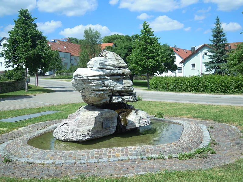 Brunnen in Rehau Brunnen mit großen weißen Steinen und Wasser, umgeben von kreisförmig gepflastertem Weg. Im Hintergrund sind Bäume vor Wohnhäuser bei blauem Himmel.
