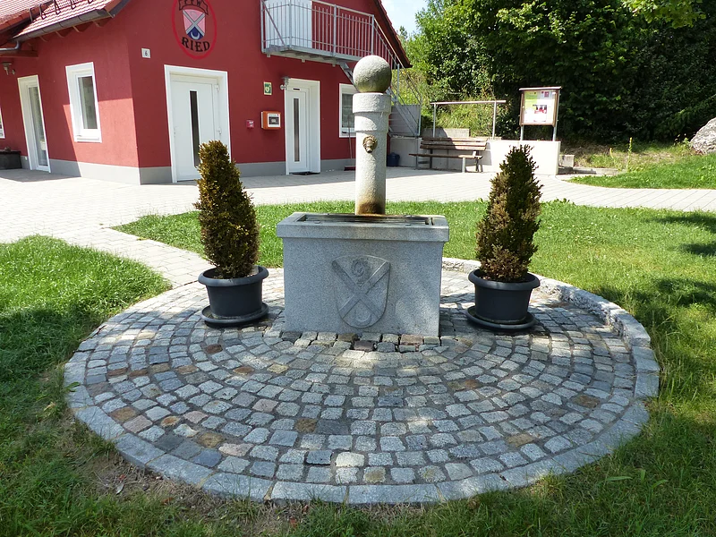 Steinbrunnen mit eingearbeitetem Wappen und zwei Pflanzen in Töpfen auf gepflastertem Rundplatz vor rotem Gebäude mit weißen Türen und Fenstern, umgeben von grüner Wiese.