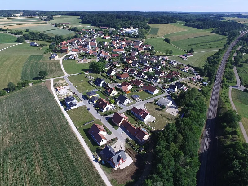Blick über Weilheim Luftaufnahme eines Dorfes mit Feldern, Bäumen, Wohnhäusern und einer Eisenbahnstrecke, die rechts im Bild verläuft.