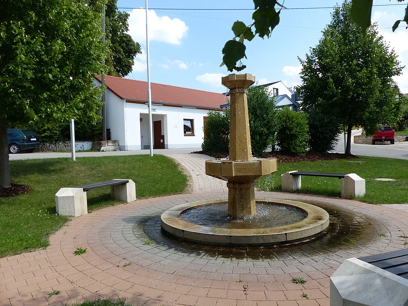 Brunnen in Weilheim Im Vordergrund ein steinerner Brunnen mit Wasser, auf gepflastertem Platz, umgeben von mehreren Bänken, Bäumen und einem weißen Gebäude mit rotem Dach im Hintergrund. Strahlend blauer Himmel mit vereinzelten Wolken.