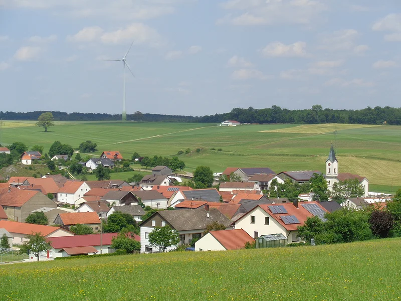 Blick über Wittesheim Dorf mit roten Dächern, einer Kirche mit Turm, grünen Wiesen und einem Windrad im Hintergrund, unter blauem Himmel.