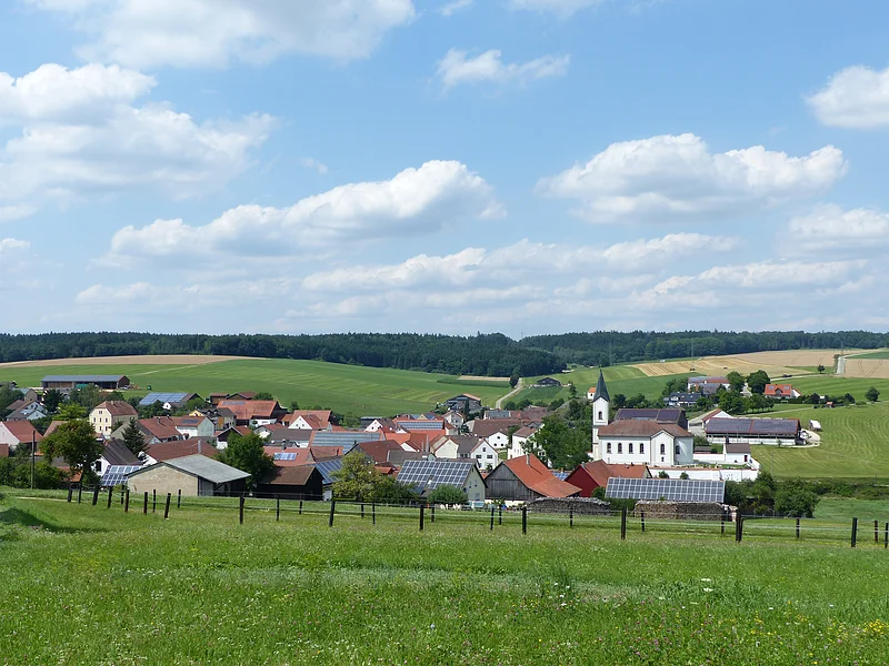 Wittesheim Aufnahme des kleines Dorfes Wittesheim mit roten Dächern und einer Kirche, umgeben von grünen Feldern und Wäldern unter blauem Himmel mit Wolken. Vorderhalb ein eingezäunter Bereich.