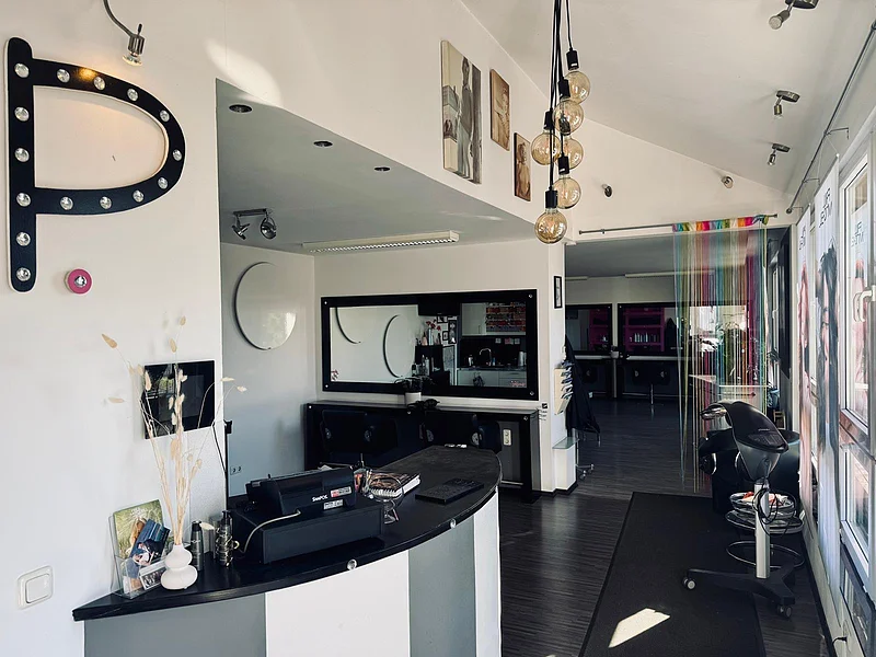 Friseursalon mit Empfangstresen, Spiegeln, Stühlen und bunten Vorhängen am Fenster im Hintergrund.