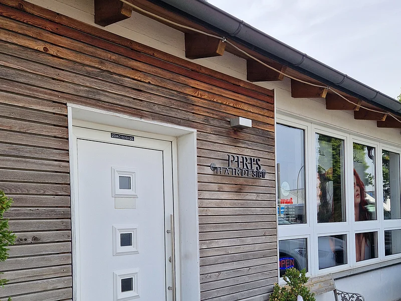 Eingang eines Friseursalons mit weißer Tür, Holzfassade, zwei Pflanzen in Töpfen und Fenster mit Werbebild.