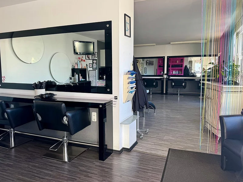 Friseursalon mit schwarzen Stühlen, großen Spiegeln, bunten Vorhängen und Pflanzen am Fenster.