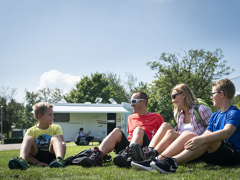 Familie am Wohnmobilstellplatz Eine Gruppe von vier Personen sitzt auf einer Wiese vor einem Wohnmobil und lacht.