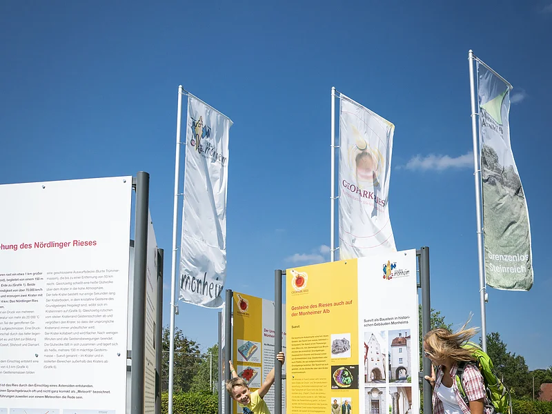 Infostelle Geopark Ries bei Monheim Ein Kind und eine Frau betrachten Informationstafeln im Freien, umgeben von Fahnen.