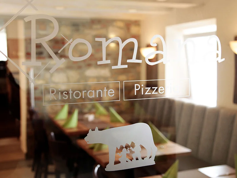 Pizzeria Ristorante Romana - unsere neuen Räume für Ihre Feierlichkeiten Fensterglas einer Pizzeria mit weißem Schriftzug „Romana Ristorante Pizzeria“ und Logo, dahinter unscharf zu sehen gedeckte Tische mit grünen Servietten und einer Steinwand.