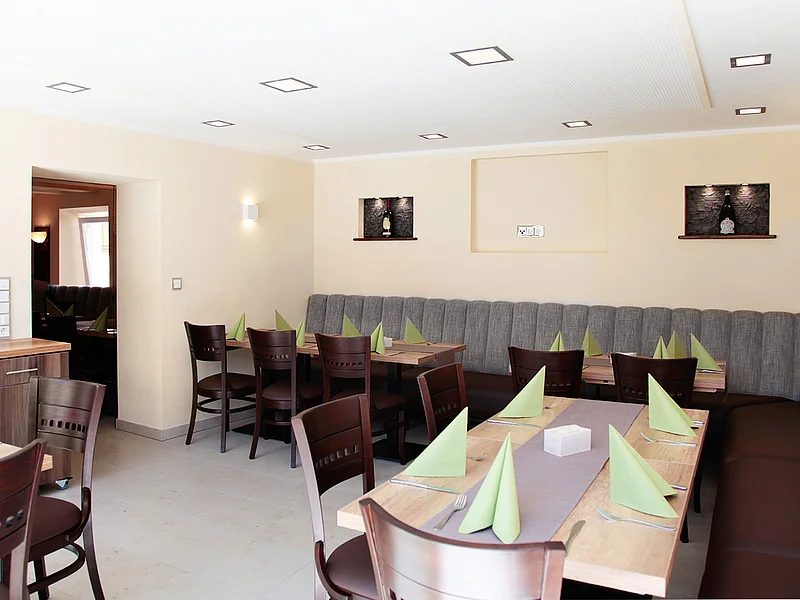 Pizzeria Ristorante Romana - unsere neuen Räume für Ihre Feierlichkeiten Blick in einen hellen Gastraum einer Wirtschaft mit Holztischen, Holzstühlen, grau-braunen Polstersitzbänken an den Wänden, grünen Servietten und Besteck. In den beige gestrichenen Wänden sind drei dekorative beleuchtete Ausfräsungen.