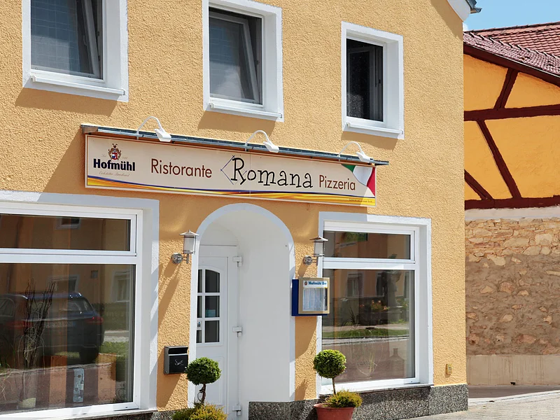 Pizzeria Ristorante Romana - Mediterraner Charme in Monheim Gelbes Gebäude mit mehreren Fenstern und einem Schild "Ristorante Romana Pizzeria" über dem Eingang. Zwei kleine Bäume links und rechts vor der Tür.