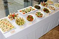 Pizzeria Ristorante Romana - wir bieten Partyservice & Catering Buffet mit vielen verschiedenen Salaten und Antipasti mit kleinen Schildern daneben auf einem weiß gedecktem Tisch vor einem Glasfenster.