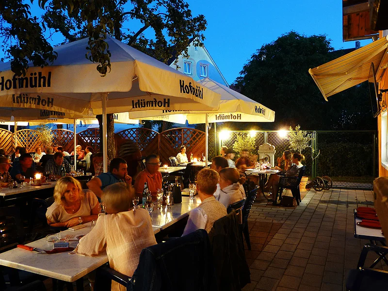 Pizzeria Ristorante Romana - große Terrasse für schöne Stunden im Freien Abendliche Außengastronomie mit Gästen an Tischen unter großen Sonnenschirmen und Beleuchtung auf gepflastertem Boden.