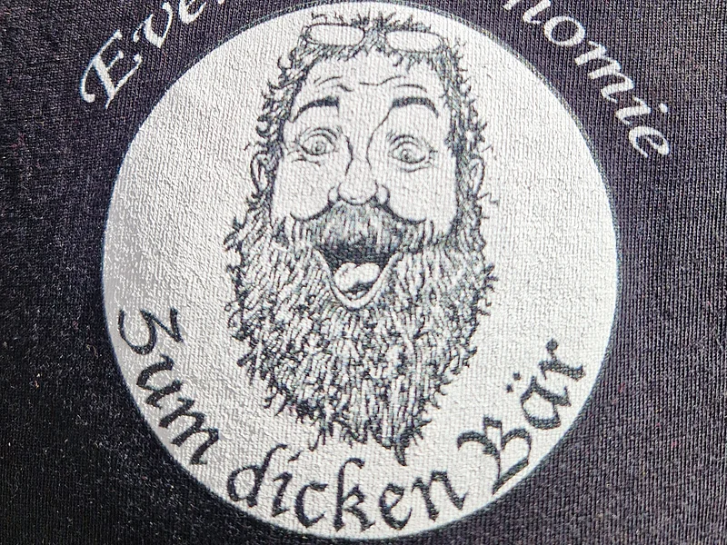 Logo zur Eventgastronomie "Zum dicken Bär" Logo mit einem Foto eines lachendem Mannes mit langem Bart und Brille. Darüber der Schriftzug „Eventgastronomie und unterhalb die Schrift "Zum dicken Bär“ auf dunklem Stoff.