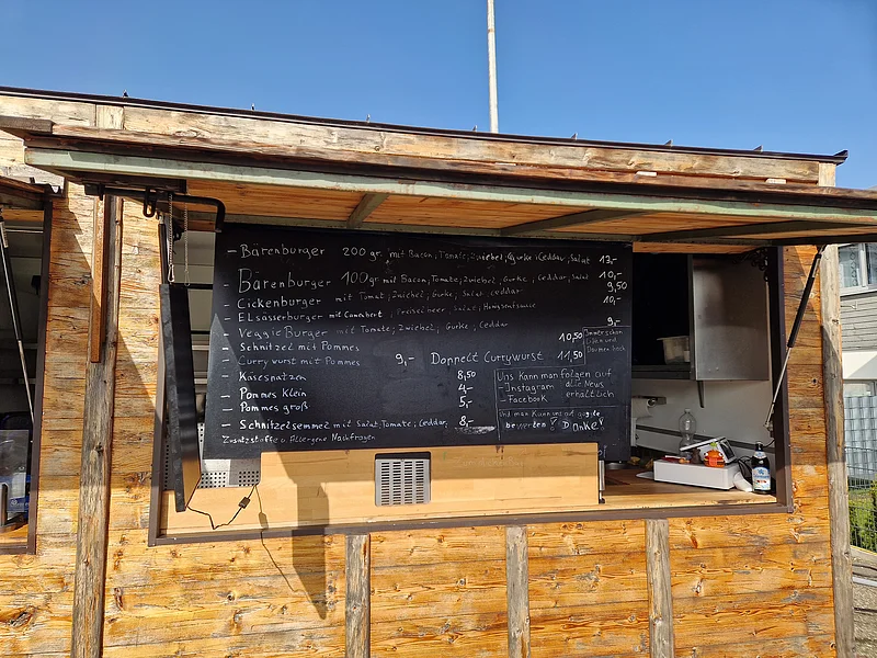 Speisekarte vom Imbiss "Zum dicken Bär" in Monheim Foto einer schwarzen Speisekarte mit Burgern, Pommes, Currywurst und Preisen an einem Holzimbiss bei Sonnenschein.