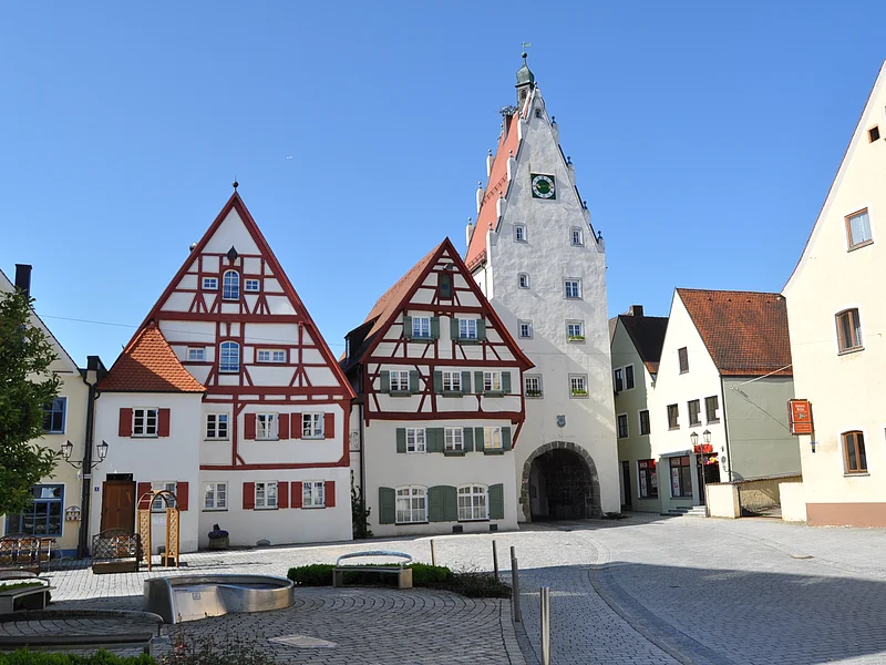 Moserhäuser mit dem Oberen Torturm Historische Fachwerkhäuser in einer Innenstadt mit einem weißen Uhrenturm an einem gepflasterten Platz bei klarem Himmel. Im Vordergrund ist ein Brunnen zu sehen, umgeben von drei Sitzbänken.