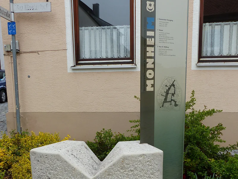 Buchstabenweg M-O-N-H-E-I-M Steinerner grauer Buchstabe M vor Infotafel aus Glas mit Erklärtext und einem Stadtplan. Auf der Infotafel vertikal der Text „Monheim“. Im Hintergrund ein Haus in beige mit einer Hecke und Straßenschildern.