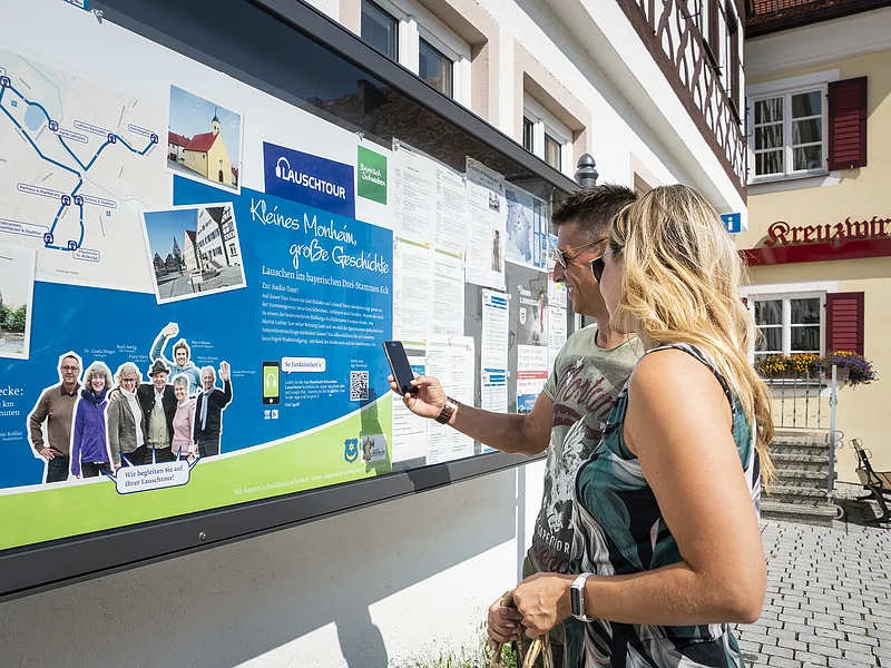 Zwei Personen vor einem weißen Gebäude, der Touristinformation Monheim betrachten eine Informationstafel mit einer Karte und Texten zur Lauschtour in Monheim und haben ein Smartphone in der Hand, mit dem sie einen Code abscannen.