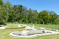 Rundes Kneippbecken in Monheim mit einem Geländer und einem Barfußpfad auf einer Wiese, umgeben von einer großen Grünfläche und Bäumen und Sitzmöglichkeiten. Ein Zaun im Hintergrund.