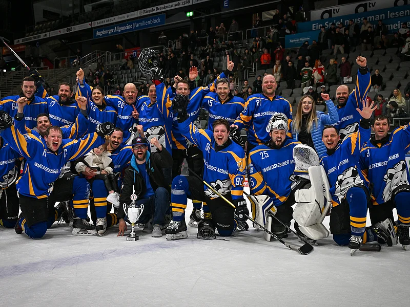 Eishockeyteam in blauen Trikots kniet auf Eis vor Zuschauerrängen, jubelt mit Pokal vor sich.