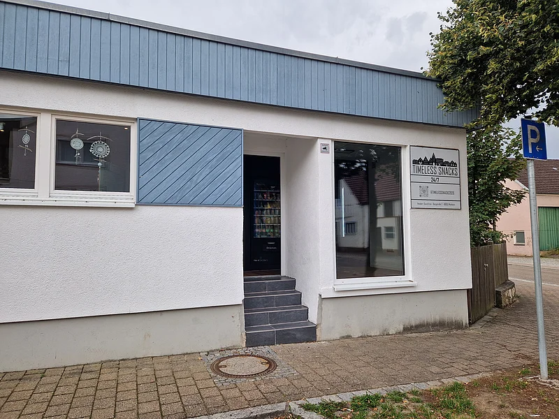 Eingang eines weißen Gebäudes mit blauen Holzverkleidungen und einem Schaufenster. Rechts vom Schaufenster ein Schild mit der Aufschrift „Timeless Snacks 24/7“ neben einer Treppe.