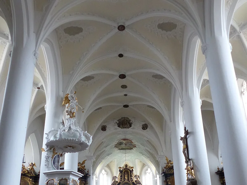 Stadtpfarrkirche St. Walburga Monheim - Blick über den Hauptgang zum Altarraum Innenansicht einer Kirche, der Stadtpfarrkirche St. Walburga in Monheim, mit hohen weißen Säulen, Gewölbedecke, Emporen mit Verzierungen und hölzernen Kirchenbänken auf beiden Seiten. Ein Altar im Hintergrund.