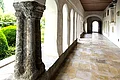 Kreuzgang des ehemaligen Klosters Monheim Blick in einen langen Kreuzgang mit Säulen und Rundbögen links im Bild, dahinter grüne Büsche. Rechts im Bild weiße Wand mit einer Ausfräsung in der Wand und Steinplatten mit Text angebracht. Entlang des Gangs alter Bodenbelag aus hellen alten Steinen.