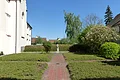 Der Klostergarten Monheim Ehemaliger Klostergarten in Monheim mit gepflastertem Weg in der Mitte, niedrigen Hecken, Büschen und Bäumen. Am Ende des Weges eine Statue vor einer Hecke unter blauem Himmel. Links im Bild ein weißes Gebäude.