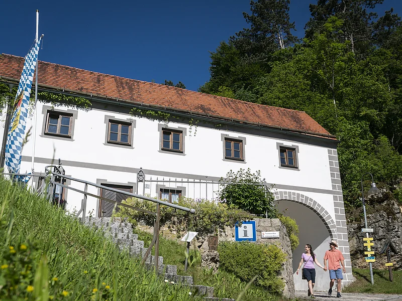 Torbogenhaus in Wellheim Weißes Gebäude mit rotem Ziegeldach, bayerischer Flagge und zwei Personen vor einem Torbogen bei Sonnenschein