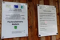 Fischereilehrhütte Monheim Zwei Informationsschilder befestigt an einer Holzwand zur Fischereilehrhütte Monheim mit Förderhinweisen und Nutzungsregeln der Fischereilehrhütte.